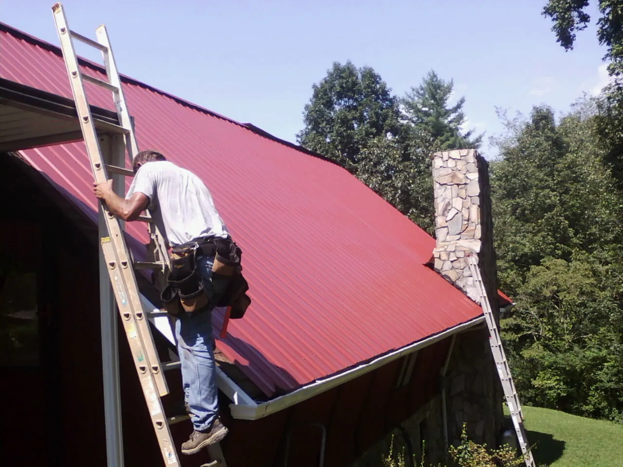 Roofer on a ladder beside a red metal roof and stone chimney — Soffit & Fascia Repair by Roofing Brothers in Bryn Mawr